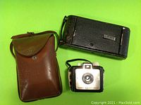 Overall view of three Kodak cameras with black expandable camera, brown leather case, and Brownie Bullet Camera.