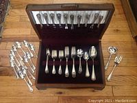 Open wooden chest with silver flatware displayed in slots and additional pieces on floor