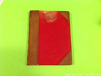 Front view of the book showing red covers with brown leather spine and corners, visible wear and a stain on front top.