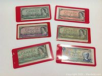 Six Canadian banknotes displayed, all with Queen Elizabeth II portrait except the $5 note with Sir Wilfrid Laurier, in red protective sleeves.