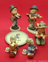 Group photo of six Hummel music motif figurines and a round plaque on a red background showing details and scale.