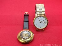 Two wristwatches placed side by side on red fabric background, showing front faces. Accutron on right with gold-tone dial and silver-tone bracelet, Tissot on left with rounded gold-tone case and leather strap.