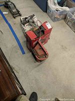 Senco RoofPro 455XP roofing nailer in red and black with attached air hose, sitting on concrete floor with box and other items nearby.