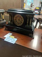 Front view of black adamantine wood Seth Thomas mantel clock with faux marble columns and decorative feet on a wooden table.