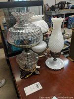 Three vintage table lamps including two milk glass lamps and one clear hurricane lamp shown grouped on a table.