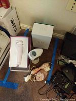 Wide view of lot showing Tria Beauty laser hair remover in box, Mia electric face brush, and cords on floor.