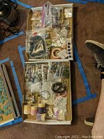 Top view of two open boxes containing assorted jewelry making supplies such as beads, buttons, cords, and metal findings spread in plastic bags and containers.