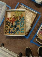 Box containing assorted colorful hand-blown glass pendants of various shapes including hearts and stars, displayed on cardboard with bubble wrap beside.