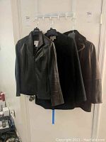 Three jackets hanging on hooks on wall. Left most is black leather jacket, middle is black fleece jacket, right is brown leather-like jacket.