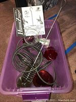 Two glass hummingbird feeders parts and assembly instructions in a purple bin, showing red glass flowers, clear glass reservoirs, and black metal hanging rods.