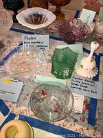 Various Fenton glass items arranged on a table with blue tape labeling, showing a pink hand-painted opalescent compote, green frosted bowl, white bell with violet decoration, and other small glass pieces.