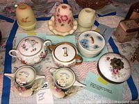 Overview of lot items showing porcelain trinket boxes, cream and sugar set, and fairy lamps on quilted cloth.