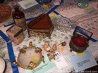Photo showing porcelain bears figurines, heart-shaped trinket box, antique trinket dish with gold accents, vintage bottle and tin, and glass pieces arranged on a cloth surface.
