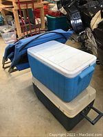 Photo shows two coolers stacked with two folded blue beach chairs leaning against them in a garage setting.