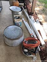 Full view showing galvanized tub, metal trashcan, PVC pipes, extension cords, and bucket of plumbing fittings.