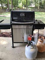 Front view of Weber propane grill with propane tank connected, showing black hood with built-in gauge and stainless steel cabinet, side shelves, and wheels.