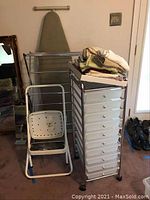 Photo showing foldable drying rack, ironing board behind it, white foldable stepstool with black grips, rolling storage drawers with linens on top, and a mirror on left.
