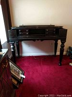Full frontal view of black walnut spinet desk on red carpet, showing top section closed with decorative panels and turned legs.
