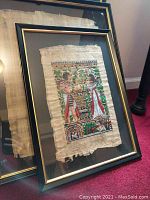 Photo of framed papyrus painting with Egyptian motifs showing two figures in robes with red and green detailing under glass in black and gold frame