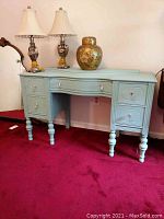 Front view of the refurbished antique dressing table painted light blue, showing five drawers and turned legs. Two table lamps and a ceramic jar placed on top.
