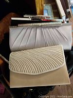 Close-up image showing two elegant clutch purses; one with fabric pleating and another with textured bead-like surface in beige/cream color.