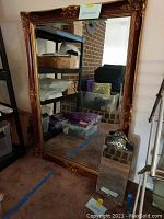 Large rectangular mirror with ornate gold gilt frame showing detailed scrollwork and floral pattern, landscape orientation, and smaller rectangular antique mirror in front with decorative top edge.