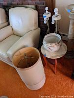 Photo showing three lamps without shades near a beige armchair, one small round wooden table, and a large ceramic cache pot