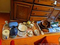 Overview of the lot showing wooden bowls, ceramic cups and saucers, vases, pitchers, and placemats laid out on plastic trays and carpet.
