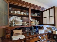 Wide view of wooden hutch with china cabinet open, showing Wedgwood Charnwood dishes, glassware on lower shelf including compotes and decanters, and stacked plates.