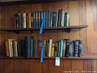 Two shelves filled with mostly hard cover books including fiction and reference titles. Various colors and sizes. Shelf and wall background visible.