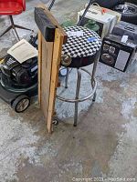 Side view of a black metal stool with a black-and-white checkered round padded seat and a wooden mechanic creeper leaning beside it on a concrete floor with other workshop items in the background.