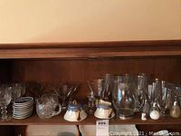 Shelf with assorted glassware including clear drinking glasses, crystal stemware, Bavaria sugar and creamer set, salt and pepper shakers, and decorative picture with silver base