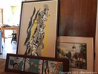 Nine pieces of framed wall art, including paintings, needlework, and prints, arranged against a wooden wall.