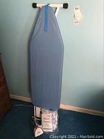 Full view of the ironing board standing upright against a blue wall, showing the blue cover and white frame. The Black and Decker iron and manual are visible at the base.