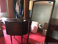 Photo showing the wooden occasional table with three drawers and the full-length mirror standing on red carpet in a room with some clutter.