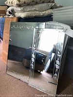 Two mirrors leaning against a wall, one large with arched top and etched floral patterns, the other smaller with etched floral border. Reflective surfaces and signs of wear visible.