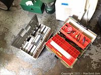 Two open fishing tackle boxes showing compartments and some accessories on a concrete floor.