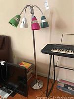 Full view of the floor lamp showing the green, white, magenta, and teal plastic shades attached on flexible chrome arms with a round weighted base. Situated beside a keyboard and other household items.