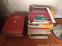 Stack of books including 9 red hardcover Scribner Radio Music Library volumes and a pile of softcover sheet music books on carpet