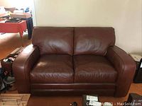 Front view of brown leather two-seater sofa showing scratches and wear on the cushions and arms.