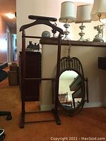 Wooden valet stand and wooden oval mirror leaning against a wall, showing full view of both items.