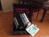Accordion inside vintage suitcase with accordion sheet music books beside it on orange carpet.