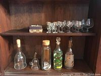 Photo showing various glassware on wooden shelves including four glass decanters with different stoppers and decorative designs plus a set of smaller glasses and shot glasses