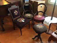 Three vintage wooden chairs with floral needlepoint embroidery, each with carved wooden frames, arranged on an orange carpet near other furniture. One swivel stool with matching upholstery and metal feet positioned in front.