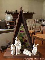 Full view of the nativity set with four white ceramic figurines standing on a wood platform in front of a triangular wood frame with a stained glass panel inside.