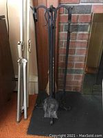 Front view of metal fireplace tool set with all tools on stand in front of brick wall and a fireplace.