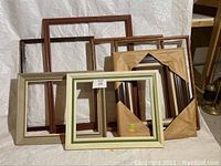 Photo shows a group of seven large wooden picture frames of assorted colors and finishes, some with smooth matte and polished wood, with one frame partially wrapped in brown paper.