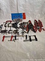 Overview of assorted clamps spread out on a white surface, showing different types and sizes including red-handled bar clamps and multiple metal C-clamps.