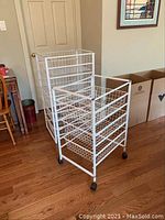 Photo showing two metal frame storage units with sliding wire baskets: one smaller unit with 3 baskets and one larger with 5 baskets both on castors on hardwood floor.