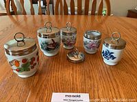 Group photo of all 5 porcelain egg coddlers with 6 metal lids displayed on wooden table.